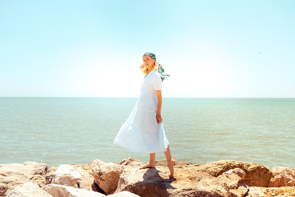 Elegant woman in a flowing white linen dress standing on seaside rocks