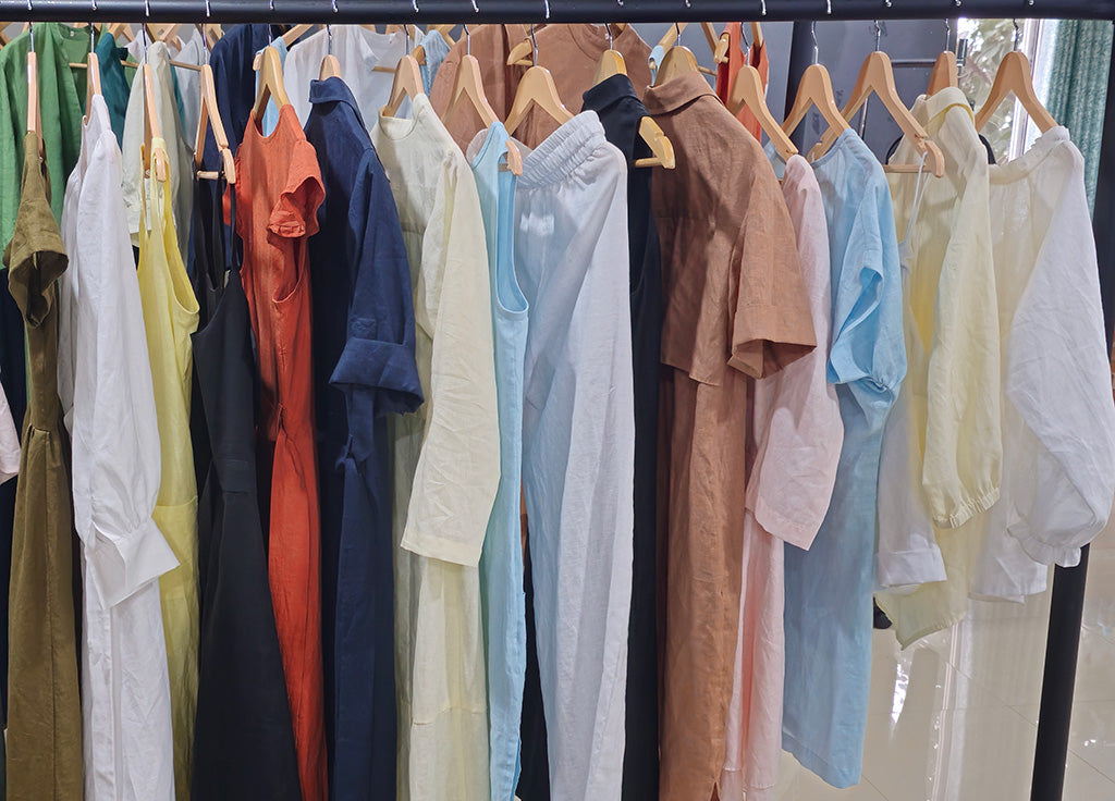 European linen dresses and shirts in multiple colors including white, navy, beige, and green, hanging on wooden hangers