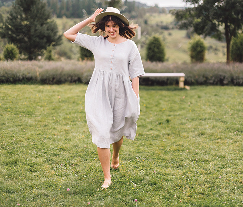 Woman in a light grey European linen dress with pockets, walking barefoot outdoors in a scenic countryside meadow