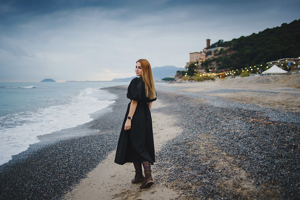 Woman wearing a black midi dress walking along a peaceful beach at dusk