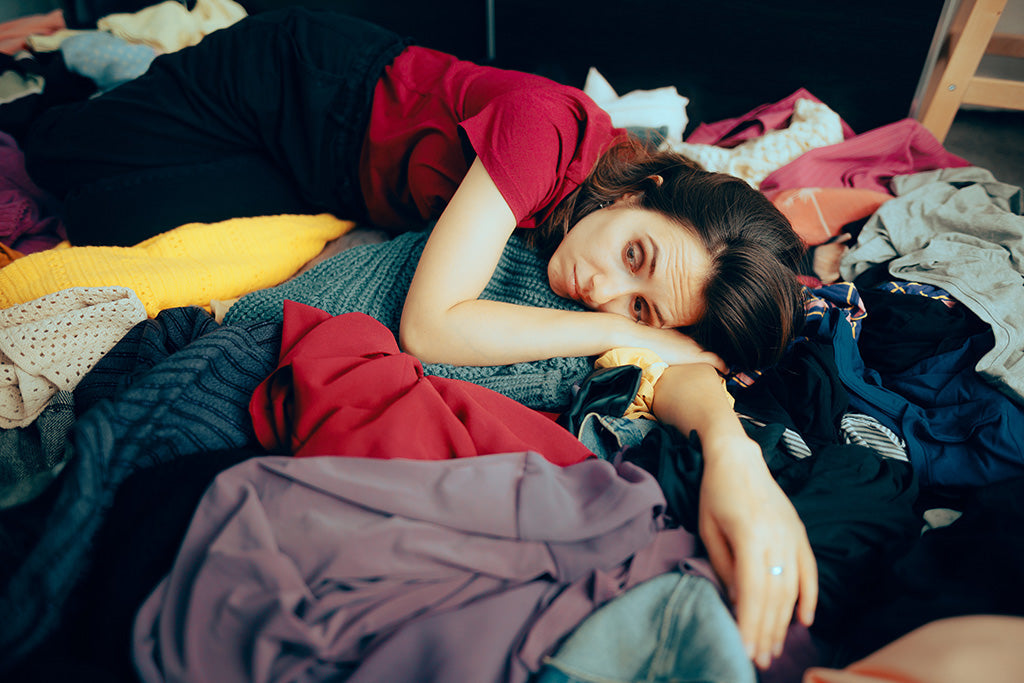 Overwhelmed woman lying on a pile of clothes, representing fast fashion clutter and unsustainable wardrobe habits