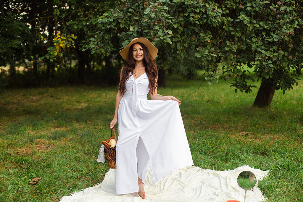 Woman wearing a long white linen dress and sun hat in an orchard