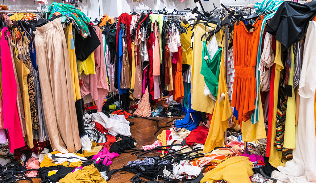Messy clothing store with colorful garments hanging on racks and piles of clothes scattered across the floor