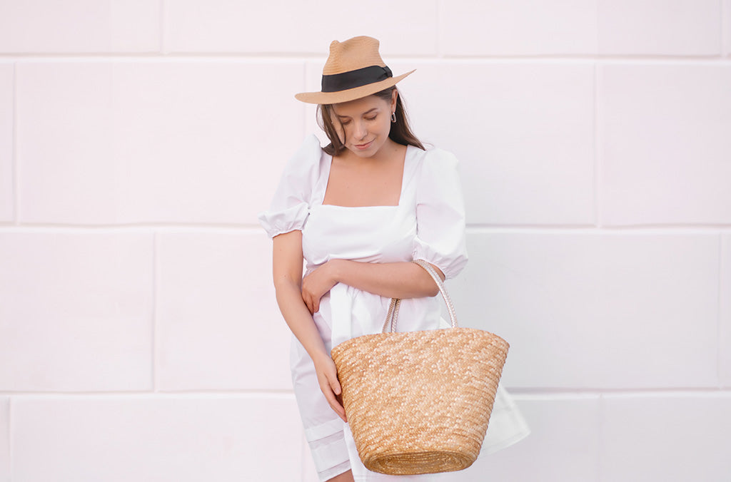 Woman wearing a white puff sleeve linen dress with a straw hat and woven tote bag