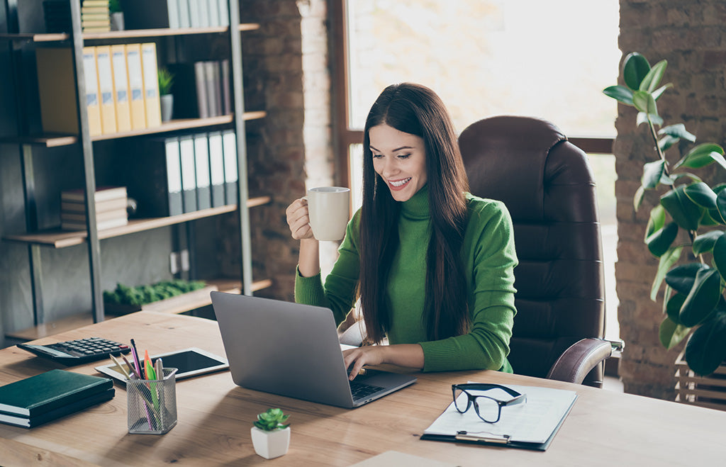 Woman in a green sweater working on a laptop while enjoying coffee in a modern office workspace
