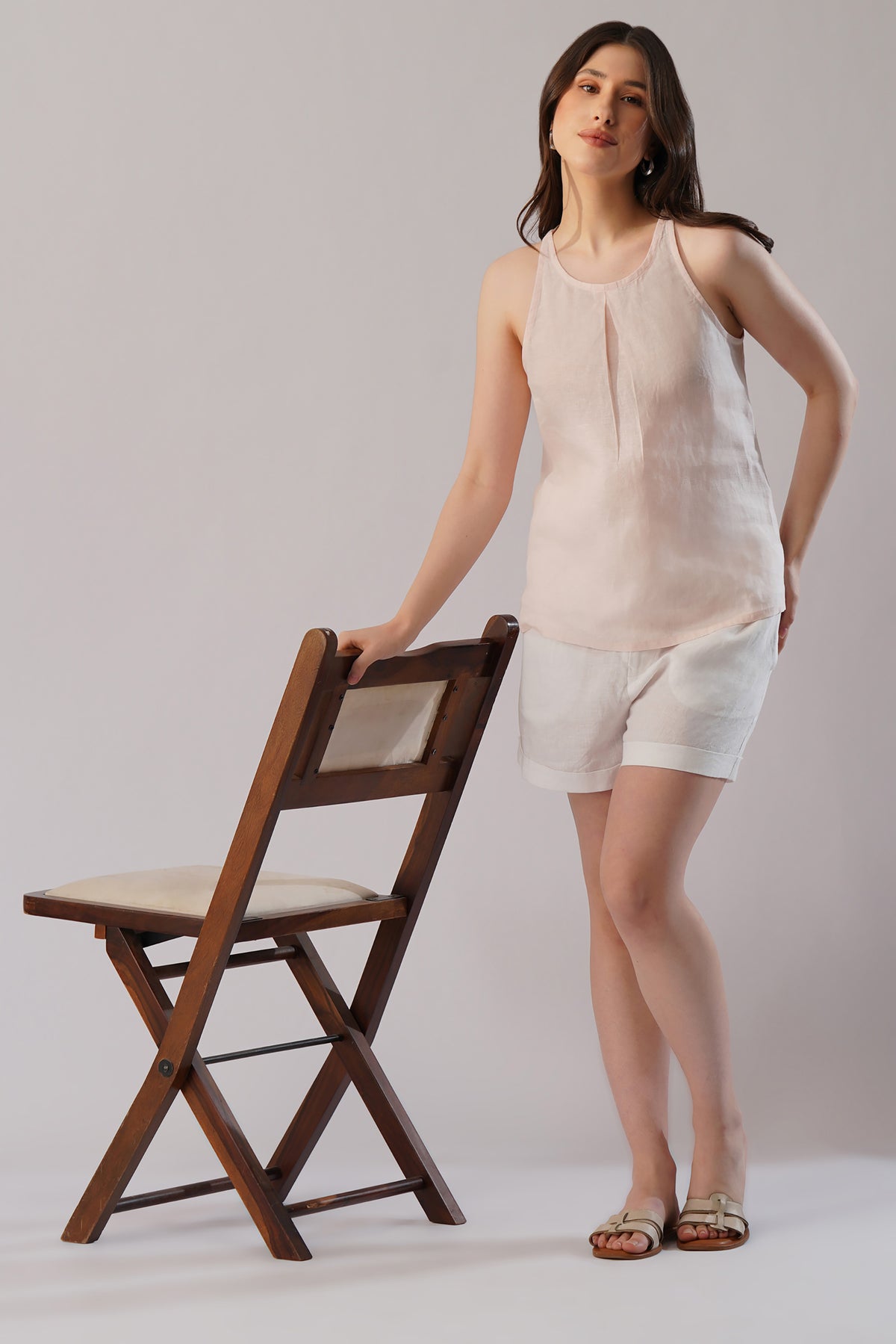 Woman in a sleeveless linen top and shorts standing next to a wooden chair 